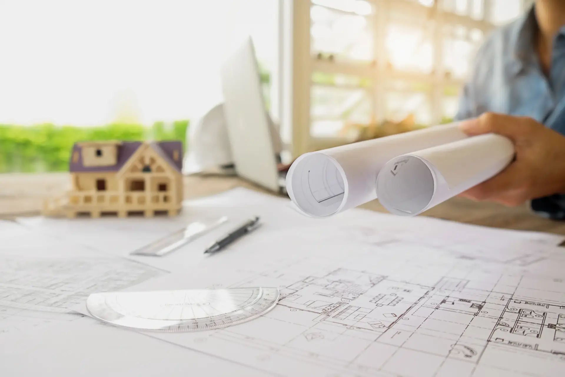Person holds architectural blueprints on a desk with a small model house by custom home builders in Anderson SC.