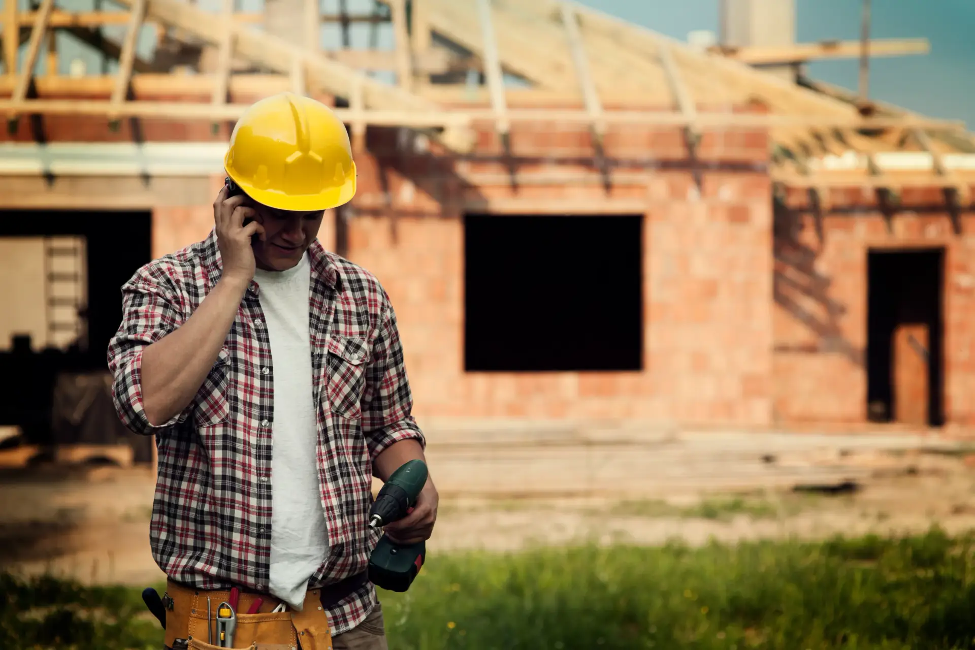 A construction worker in a yellow hard hat talks on a phone while holding a drill during Custom Home Building in South Carolina.
