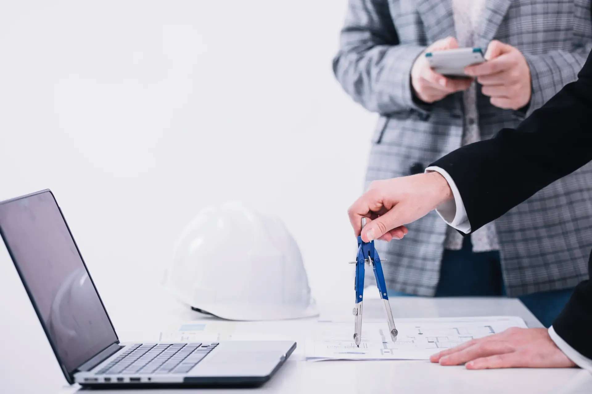 Professionals using a compass and blueprints for Construction Consulting in South Carolina near a laptop and hard hat.
