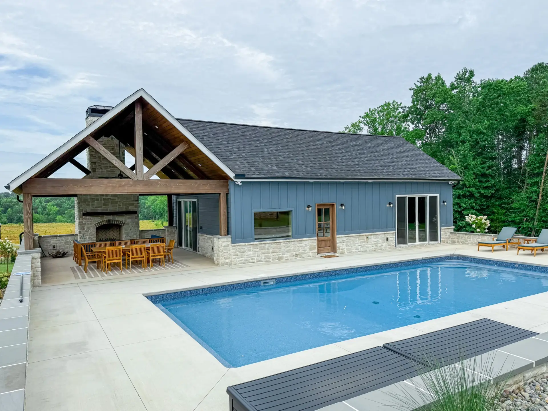 A sparkling blue swimming pool next to an upscale Custom Luxury Pool House featuring a wooden pavilion and stone accents.