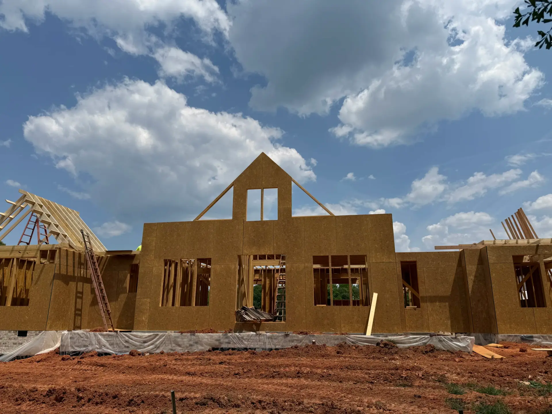 Wooden wall framing and sheathing of a Custom Luxury Farmhouse under a bright blue sky with white clouds.