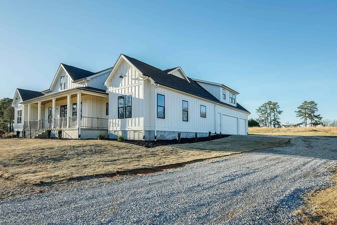Side view of a Custom Luxury Farmhouse with white board and batten siding, black windows, and a gravel driveway.